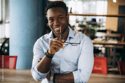 Portrait of a handsome and stylish African American guy with an attractive smile. He holds his glasses in his hand. Indoors