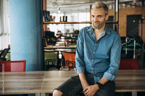 Portrait of a stylish modern guy in a blue shirt and with beard who sits in the office on the table