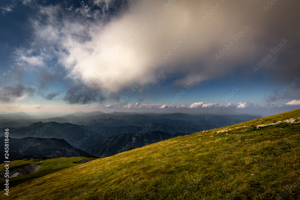 View from Rax plateau, full of fresh, green grassy meadow with blue dramatic cloudy sky to valley and alpine hills, Raxalpe, Schneeberg massif, Alps, Lower Austria