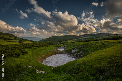 Small patch of snow in the summer in the middle of grassy meadow in rax plateau, near route Dambocksteig from Schneeberg, highest peak Kosterwappen in the cloudy sunset, Raxalpe, Lower Austria photo