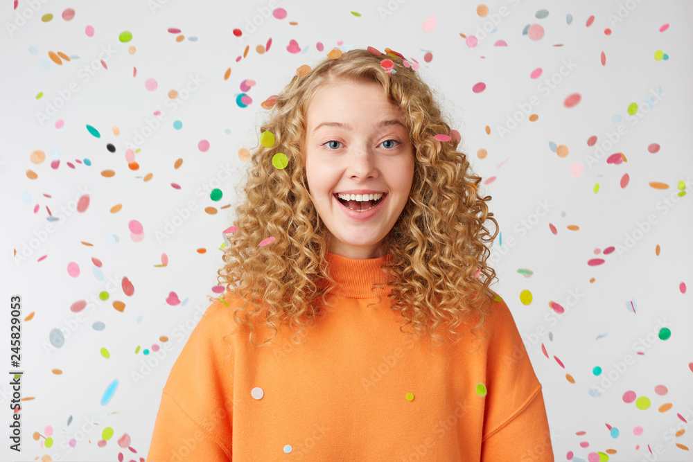 Celebrating happiness, young woman with big smile stands under the falling confetti.Curly blonde in orange sweater enjoying life moment, came to the holiday. Celebrating happiness, young woman with big smile stands under the falling confetti.Curly blonde in orange sweater enjoying life moment, came to the holiday.