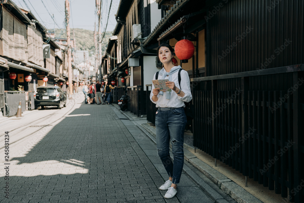 full length of young girl backpacker visiting Hanamikoji street standing on road holding paper map. japanese wooden building house hanging bamboo curtain on roof. woman finding direction destination.