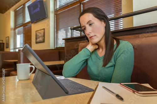 A young female works out of the office sits quietly reading the tablet in a booth. She sits alone at the cafe focused on her work at a table with a cup of coffee.