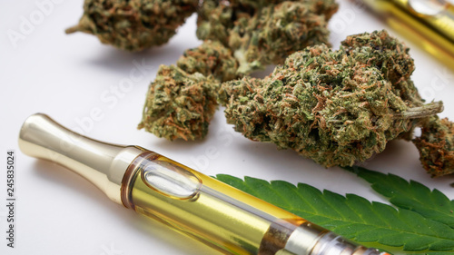 THC/CBD Concentrated Oil In Cartridge Closeup With Marijuana Herb & Glass Pipe On White