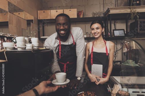 Smiling black guy in apron gives cup of cooked coffee to visitor in cafe. Confectionery. Barista.