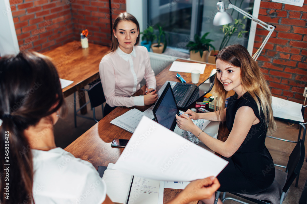 Fototapeta premium Team of business women working with papers using laptops sitting at desk in office