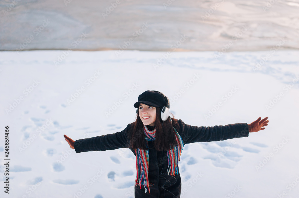 Young funny caucasian girl in headphones in winter on a snow background. Playlist with your favorite songs. To cheer up the music. Spreading hands. Copy space.