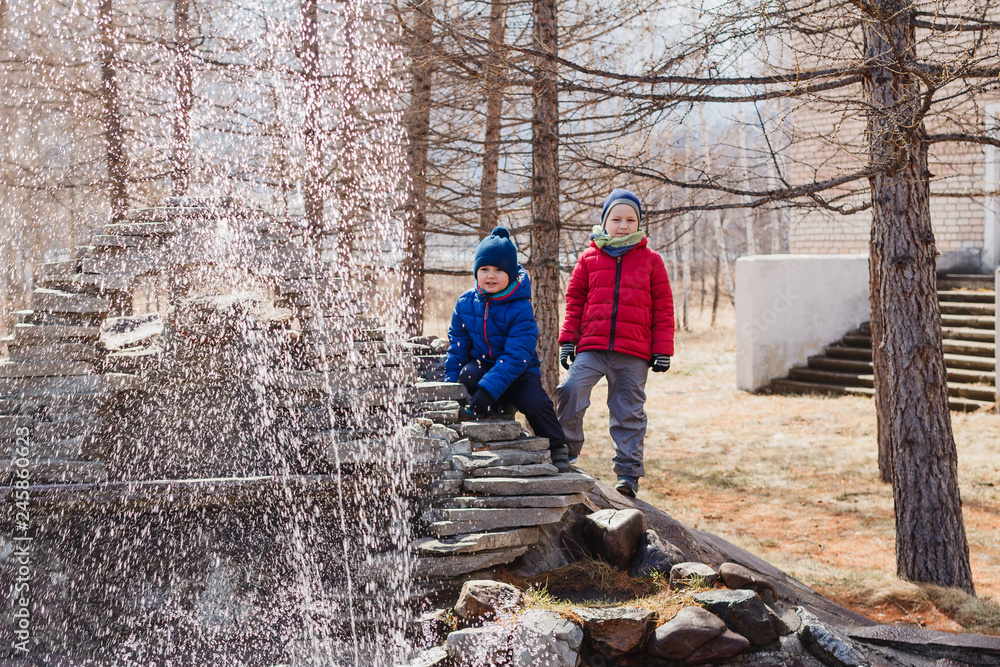 two preschooler boys play on a small fountain in the Park in early spring. children in warm clothes two preschooler boys play on a small fountain in the Park in early spring. children in warm clothes
