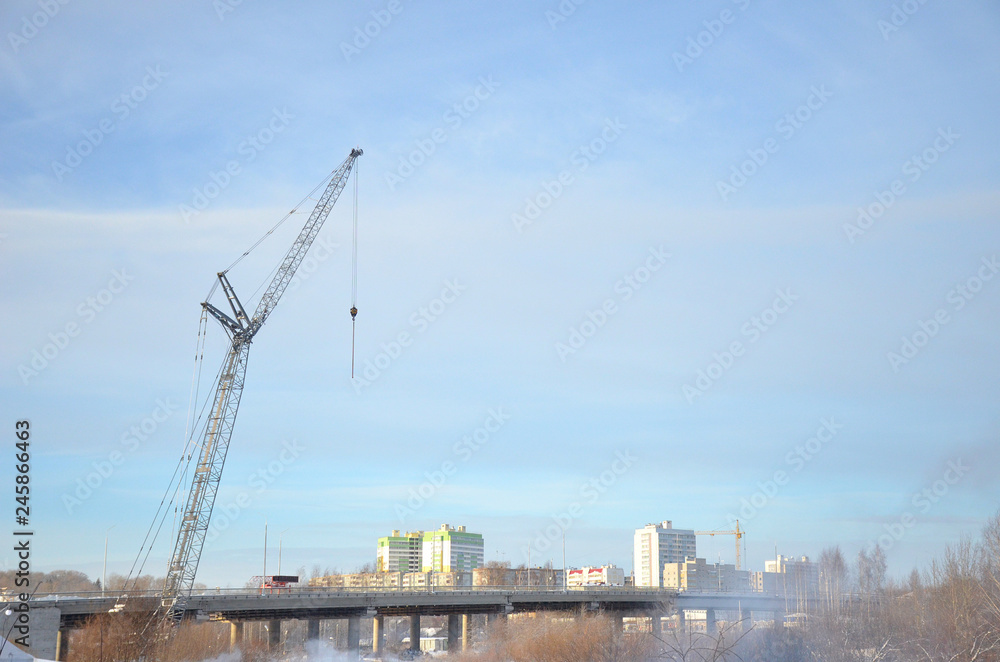 Gray tower crane towering over the bridge on the background of high-rise buildings and blue sky