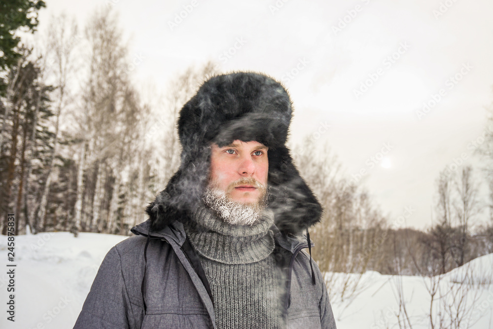 Siberian Russian man with a beard in hoarfrost in freezing cold in the winter freezes in a village in a snowdrift and wears a hat with a earflap. Siberian Russian man with a beard in hoarfrost in freezing cold in the winter freezes in a village in a snowdrift and wears a hat with a earflap.