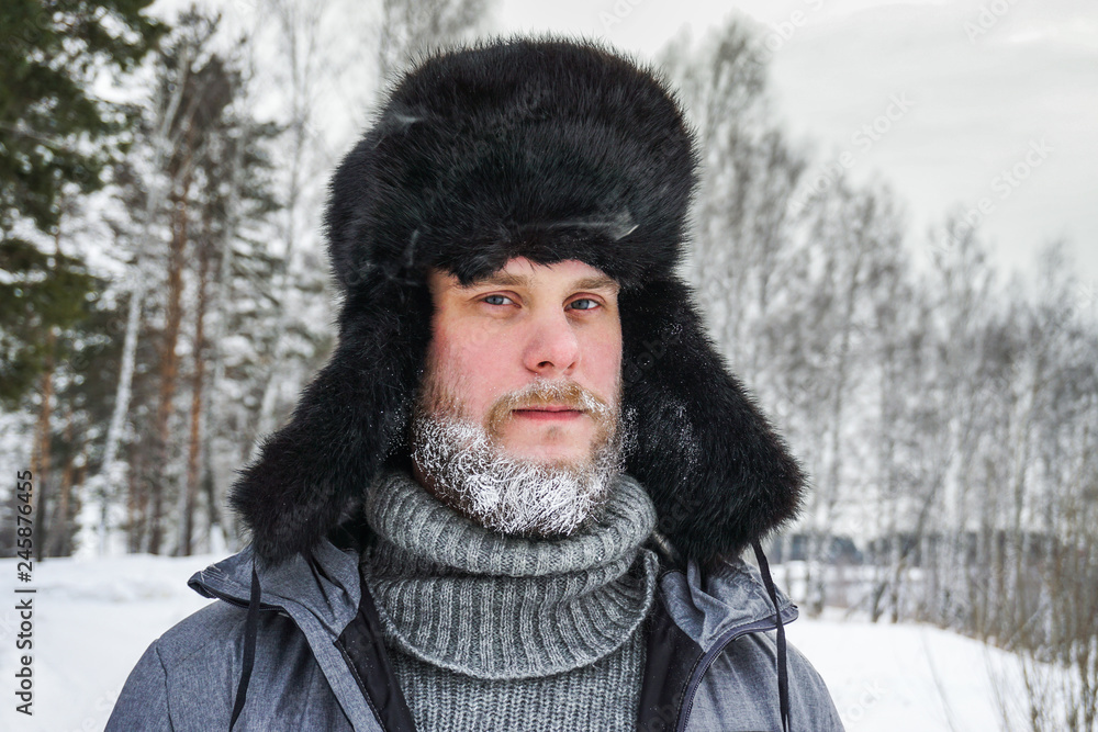 Siberian Russian man with a beard in hoarfrost in freezing cold in the winter freezes in a village in a snowdrift and wears a hat with a earflap. Siberian Russian man with a beard in hoarfrost in freezing cold in the winter freezes in a village in a snowdrift and wears a hat with a earflap.