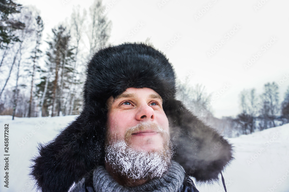 Happy Siberian Russian man with a beard smiles and lets out steam from his mouth in hoarfrost in freezing cold in the winter freezes in a village in a snowdrift and wears a hat with a earflap. Happy Siberian Russian man with a beard smiles and lets out steam from his mouth in hoarfrost in freezing cold in the winter freezes in a village in a snowdrift and wears a hat with a earflap.