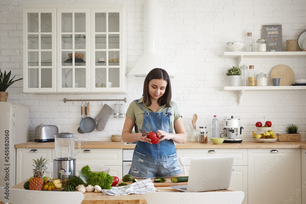 Beautiful young brunette female in denim jumpsuit cooking dinner in modern light kitchen interior, peeling red bell pepper, making healthy salad, watching video recipe online on portable computer