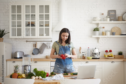 Beautiful young brunette female in denim jumpsuit cooking dinner in modern light kitchen interior, peeling red bell pepper, making healthy salad, watching video recipe online on portable computer