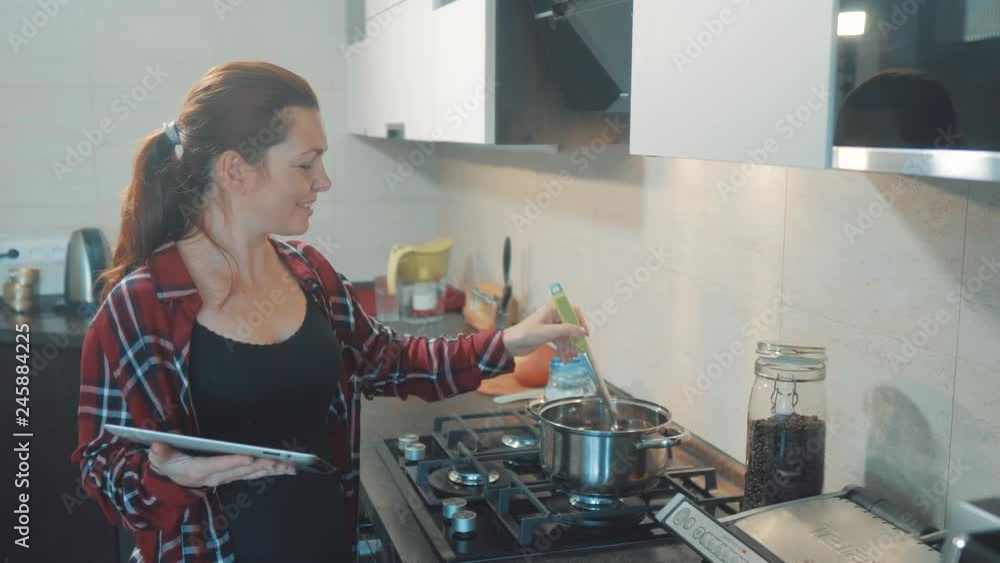 woman cooks soup pan in the kitchen prevents the water in the pot and looks at the recipe on the digital tablet. woman in the kitchen slow motion video. girl mom lifestyle in the kitchen indoors woman cooks soup pan in the kitchen prevents the water in the pot and looks at the recipe on the digital tablet. woman in the kitchen slow motion video. girl mom lifestyle in the kitchen indoors