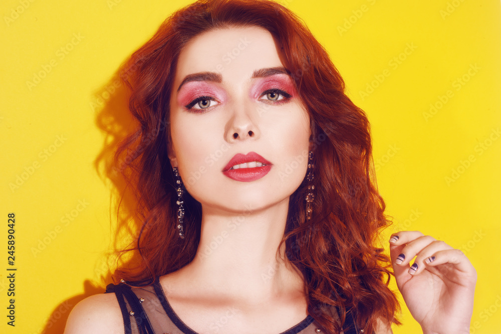 Fototapeta premium Fashion girl portrait on yellow background posing. Pretty brunette girl with long cerly hair and pink make up. Girl with long earrings. Portrait fashion model . Woman with pink make up. Beauty concept