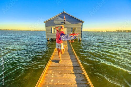 Happy caucasian woman with australian flag enjoying in front of Blue Boat House with wooden jetty on Swan River at sunset. Perth landmarks in Western Australia. Copy space.