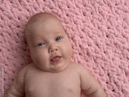 Happy and smiling baby lies and looks in the frame. The baby lies on a pink micro-polyester rug.