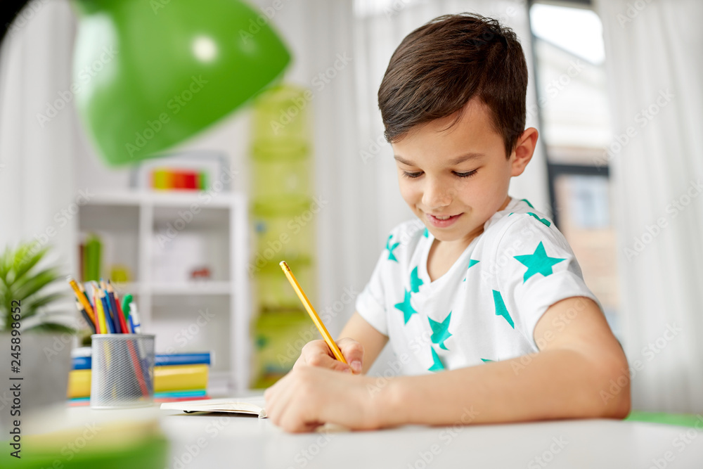 education, childhood and school concept - happy little boy sitting at desk and writing or drawing to notebook by pencil at home education, childhood and school concept - happy little boy sitting at desk and writing or drawing to notebook by pencil at home