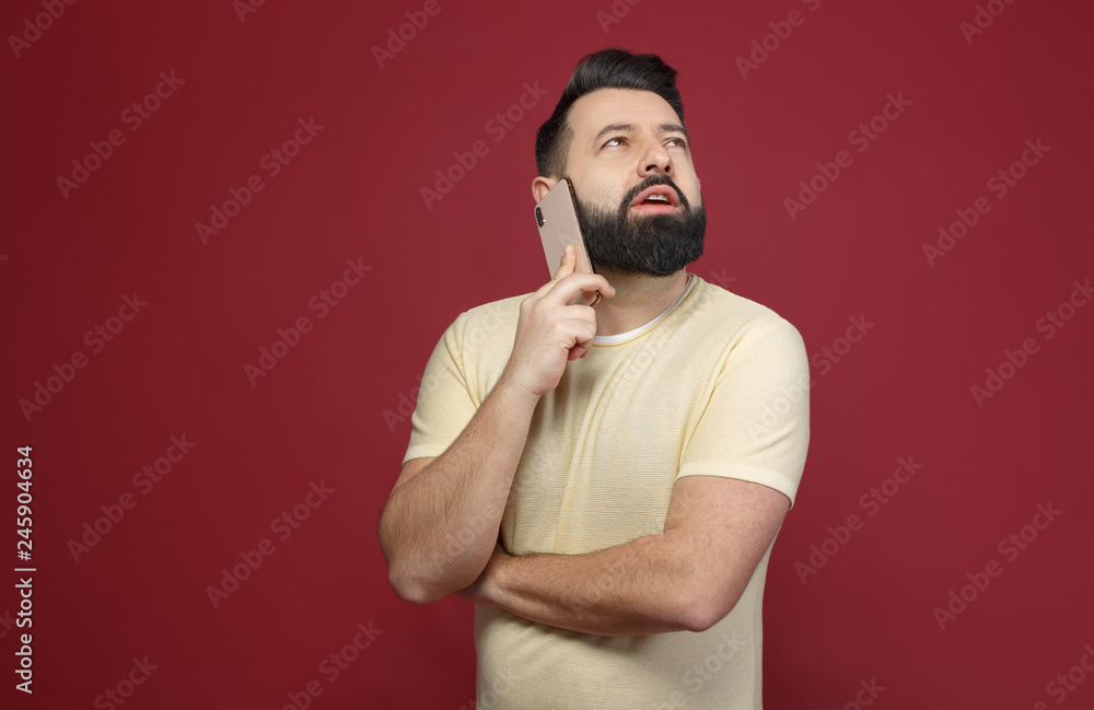 A cute and bearded brunette man on a red background says emotionally on the phone A cute and bearded brunette man on a red background says emotionally on the phone
