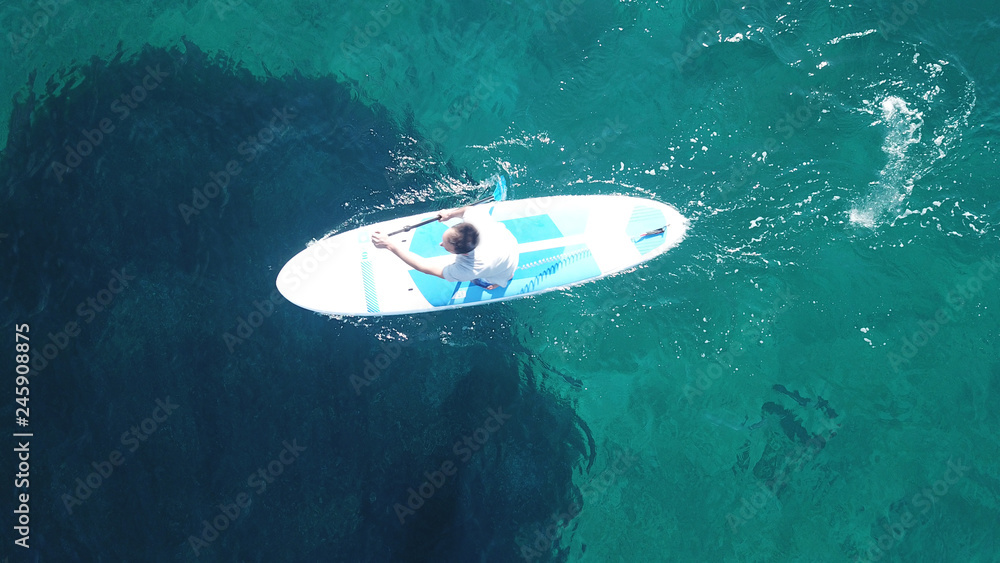 Aerial drone top view photo of young fit man exercising Stand Up Paddle surf board or sup in mediterranean turquoise sea sandy beach Aerial drone top view photo of young fit man exercising Stand Up Paddle surf board or sup in mediterranean turquoise sea sandy beach