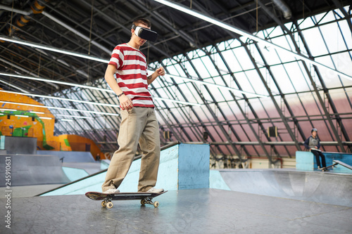 Young boy in casualwear standing on skateboard while training on parkour area or stadium and watching virtual adventure