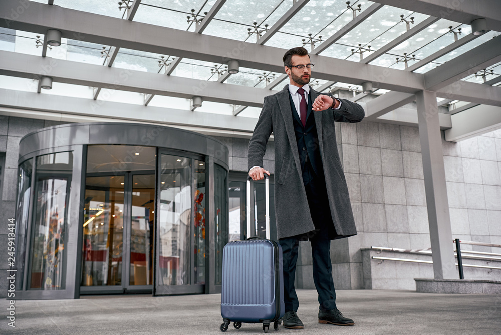 Young man checking his watch and pulling suitcase Young man checking his watch and pulling suitcase