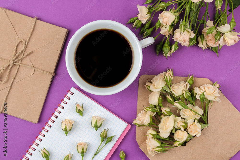 Beautiful mini roses in an envelope with a notepad and a cup of coffee on a bright lilac background. holidays. Valentine's Day. women's Day. view from above. Beautiful mini roses in an envelope with a notepad and a cup of coffee on a bright lilac background. holidays. Valentine's Day. women's Day. view from above.