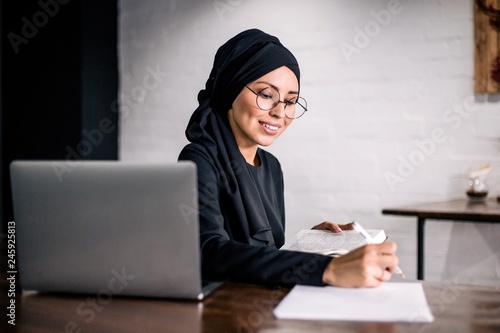 Photography Muslim girl sitting at a table with a laptop makes notes in a notebook