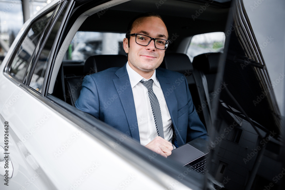 Businessman in a suit sitting in the back seat of a car working behind a laptop view from the window