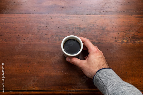Male hand holding a cup of coffee on a wooden table. Overhead view.