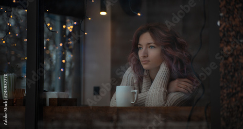 girl in a white sweater and with a white cup sits by the window in a coffee shop decorated with garlands