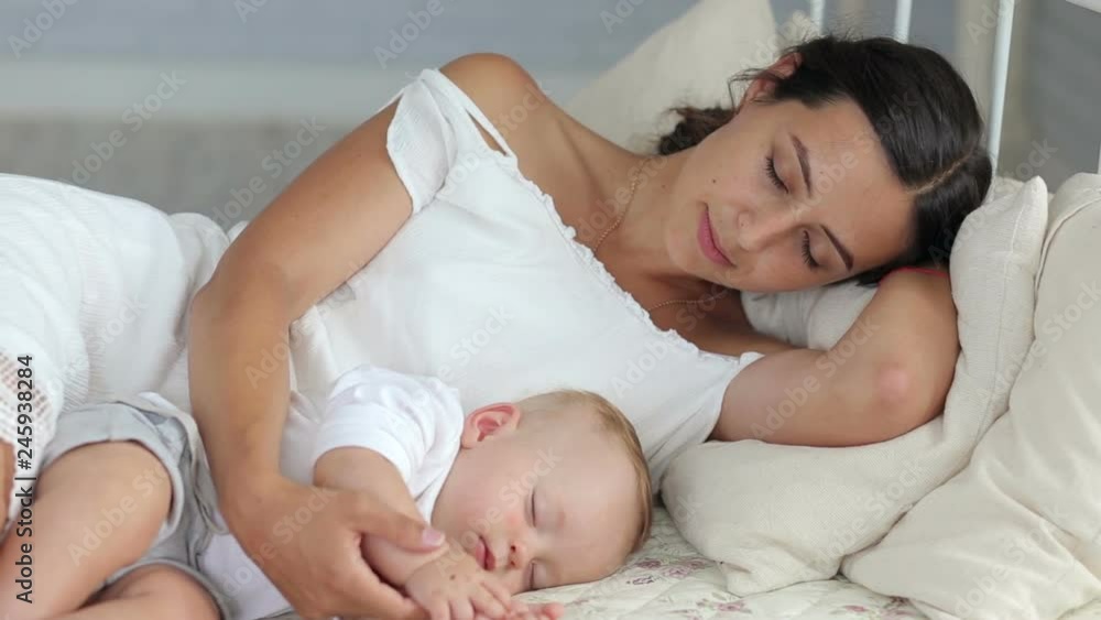 Happy mom is lying with her newborn sleeping baby in bed. Mom sleeps next to the baby. Happy mom is lying with her newborn sleeping baby in bed. Mom sleeps next to the baby.
