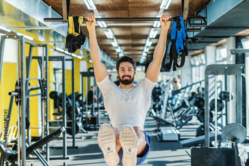 Smiling bearded muscular man in sportswear hanging from the bar and doing endurance. Gym interior.