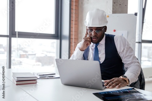 African businessman Builder in a white helmet sitting at a table in the office working on a laptop