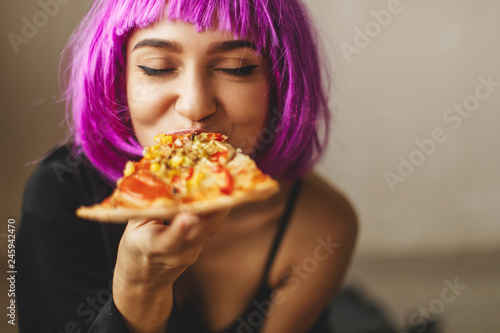 Funny pink hair wig girl in black lingerie and shirt eating pizza at home. Girl enjoying a delicious pizza. 
