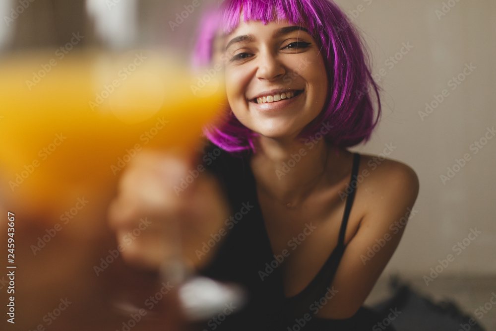 Attractive playfull woman wears pink wig, black lingerie and shirt. Girl drinking orange juice in glass. Woman sitting on the floor. She look sexy and hot, girl raised a glass. Toast