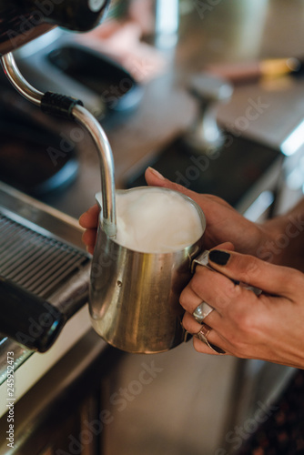 Female hands holding silver pitcher near coffee machine, barista steaming milk before adding it to espresso to make cappuccino or latte