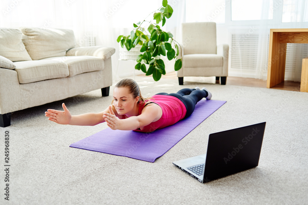 Beautiful young woman doing exercise for back on floor at home, online training on laptop computer, copy space. Full length portrait. Yoga, pilates, working out exercising Beautiful young woman doing exercise for back on floor at home, online training on laptop computer, copy space. Full length portrait. Yoga, pilates, working out exercising