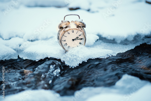 Frozen time. A clock on an ice next to the water. Extreme weather situation. Snow falling on a clock in a nature. Golden old clock. photo