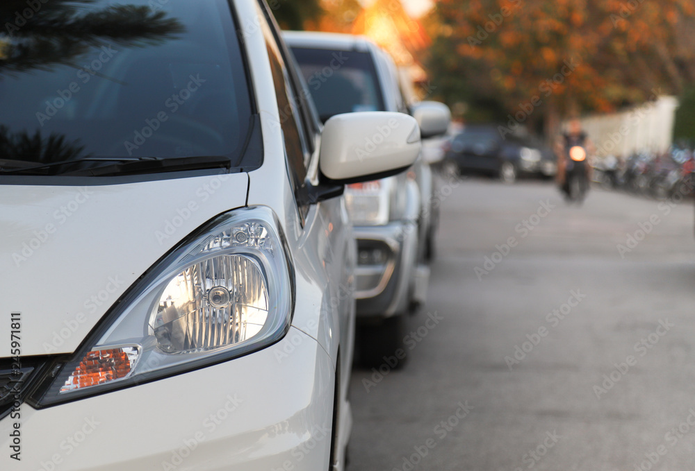 Closeup of front side of white car and other cars parking beside the street in twilight evening of sunny day with natural background. 