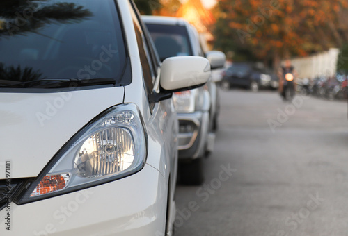 Closeup of front side of white car and other cars parking beside the street in twilight evening of sunny day with natural background.  © Amphon