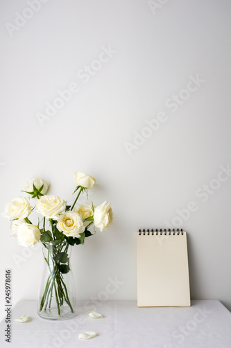 White roses in a glass vase and white blank notepad on white table top on whi...