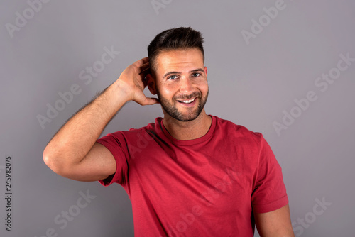 A smiling handsome young man in a red tshirt standing in front of a grey background in the studio.