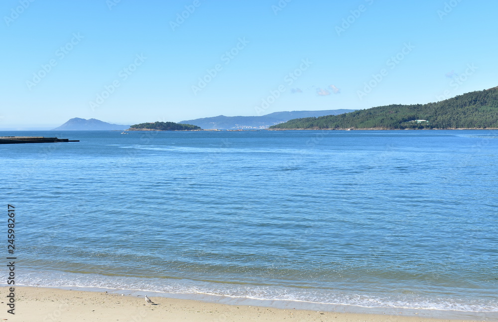 Beach in a bay with island, small coastal village and forest. Bright sand and blue sea with foam, sunny day, clear sky. Galicia, Spain. Beach in a bay with island, small coastal village and forest. Bright sand and blue sea with foam, sunny day, clear sky. Galicia, Spain.