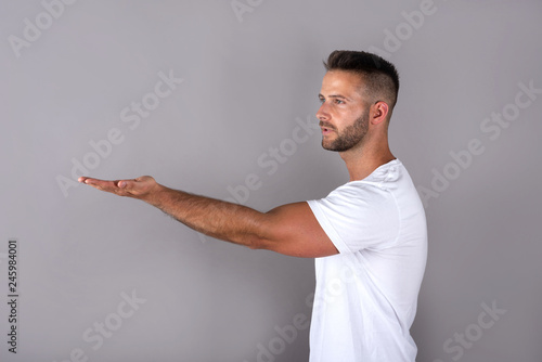 A handsome young man in a white tshirt showing something with his palms in front of a grey background in the studio.