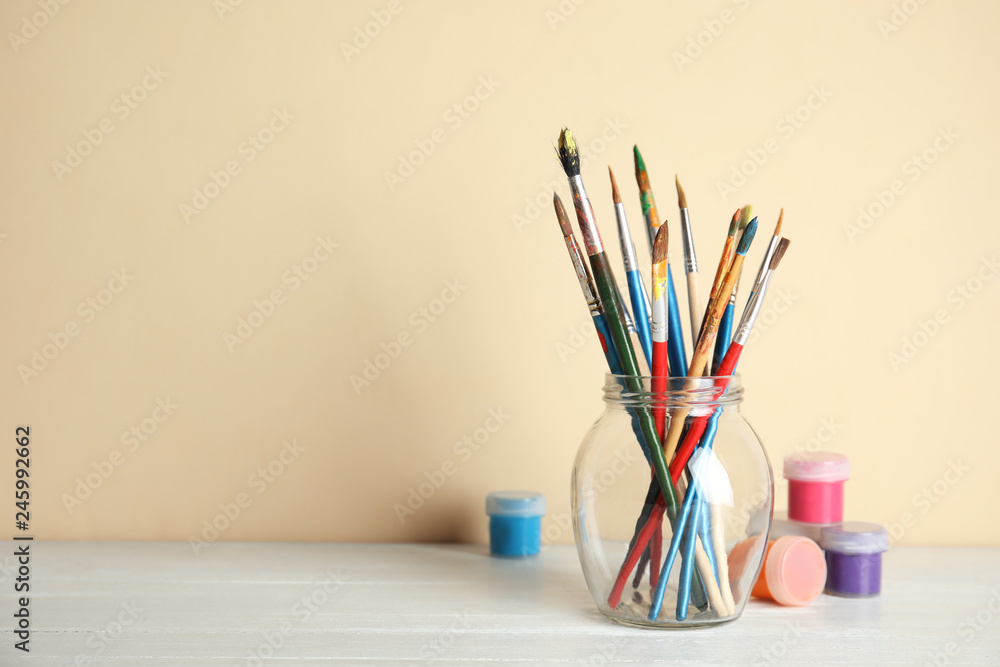 Glass with brushes and paint jars on table against color background. Space for text Glass with brushes and paint jars on table against color background. Space for text