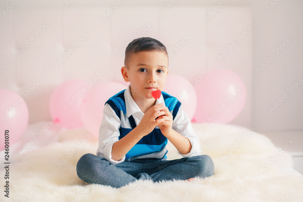 Portrait of white Caucasian cute adorable boy child eating heart shaped lollipop celebrating Valentine Day holiday. Kid eating candy sitting on bed in bedroom. Portrait of white Caucasian cute adorable boy child eating heart shaped lollipop celebrating Valentine Day holiday. Kid eating candy sitting on bed in bedroom.