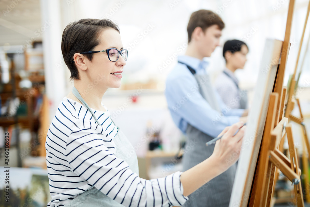 Side view portrait of smiling adult woman painting on easel enjoying work in art class, copy space Side view portrait of smiling adult woman painting on easel enjoying work in art class, copy space
