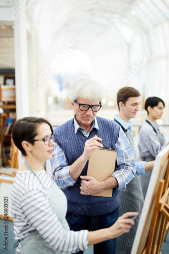 Portrait of mature art teacher helping group of students painting picture on easel in art class